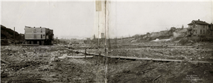 A wide muddy valley between old wooden buildings, one of them has been raised off the ground by wooden scaffolding